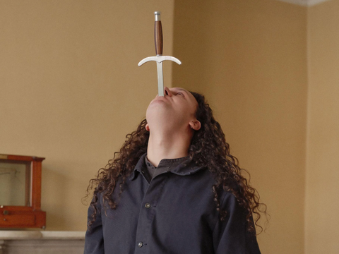 person with long curly brown hair looks to ceiling with sword in mouth