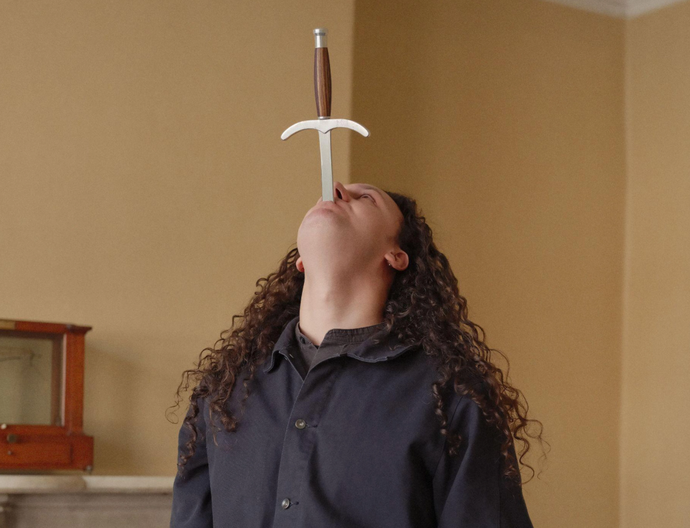 person with long curly brown hair looks to ceiling with sword in mouth