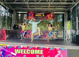 Three performers dance on an outdoor stage decorated with colorful banners reading “Camden Mela,” with trees visible behind the stage and bright traditional-style costumes in motion.
