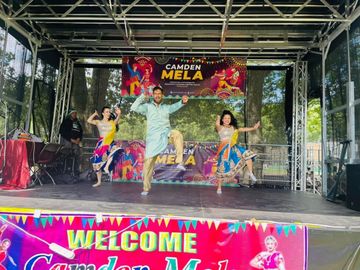 Three performers dance on an outdoor stage decorated with colorful banners reading “Camden Mela,” with trees visible behind the stage and bright traditional-style costumes in motion.
