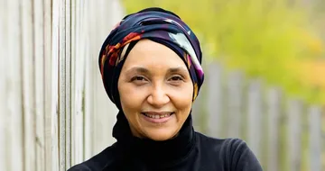Smiling person wearing a colorful headscarf and black top, standing against a wooden fence with greenery in the background.