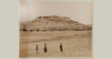 Three people walk across a field towards a large, ancient fortress on a hill under a clear sky.