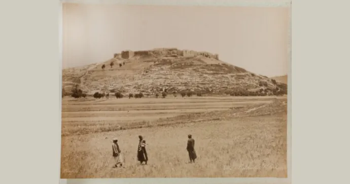Three people walk across a field towards a large, ancient fortress on a hill under a clear sky.