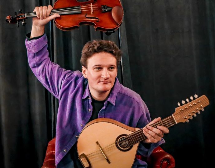 person wearing purple shirt holds two string instruments, one holding above head