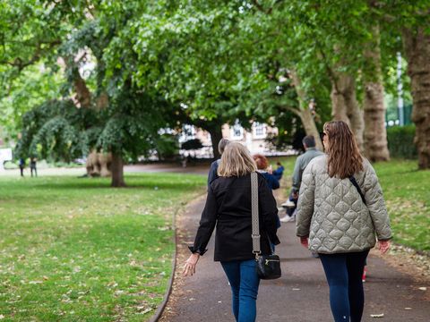 Two people walking along a park path lined with green trees on a sunny day.