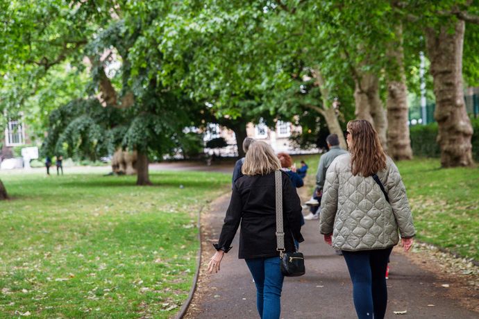 Two people walking along a park path lined with green trees on a sunny day.