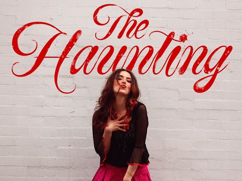 Person with long hair in a black top and bright pink skirt stands against a white brick wall, with large red script text reading 'The Haunting' above.