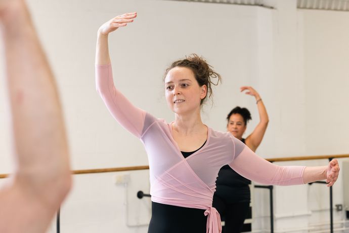 Two women practicing ballet in a studio, one in a pink wrap top, raising their arms gracefully with mirrored movements.