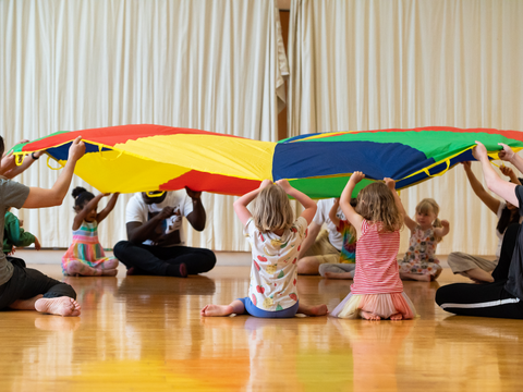 Children and adults sitting on the floor holding up a colorful parachute in a playroom with wooden floors and cream curtains.
