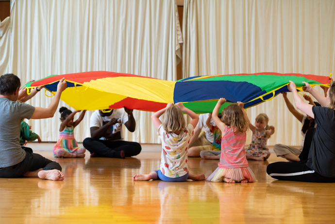 Children and adults sitting on the floor holding up a colorful parachute in a playroom with wooden floors and cream curtains.