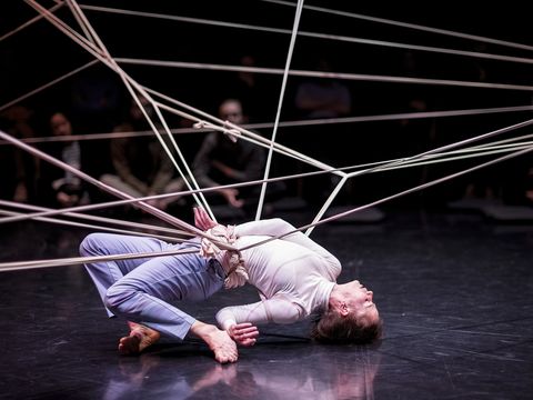 person strikes contorted pose with legs behind them and arm behind them while entangled in some silk rope which forms a spiderweb pattern above person