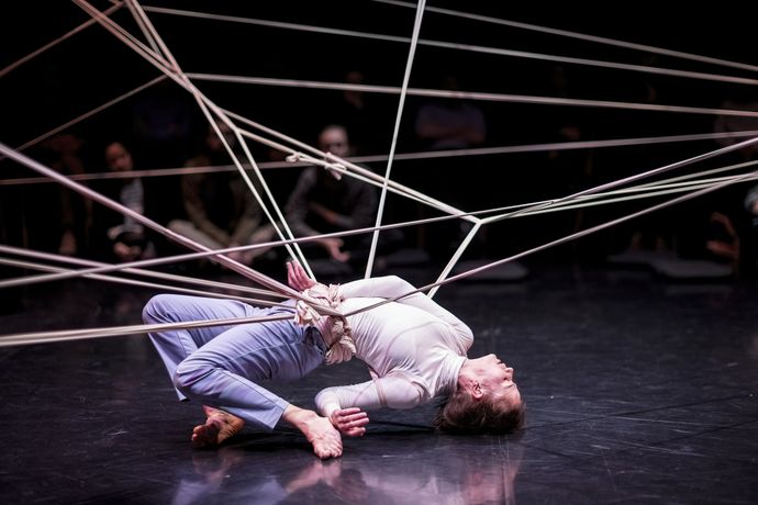 person strikes contorted pose with legs behind them and arm behind them while entangled in some silk rope which forms a spiderweb pattern above person