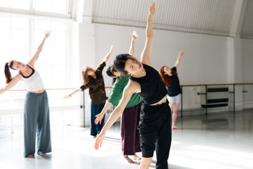People performing a dance or exercise routine in a sunlit studio, with arms extended and leaning to one side.