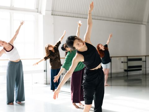 People performing a dance or exercise routine in a sunlit studio, with arms extended and leaning to one side.