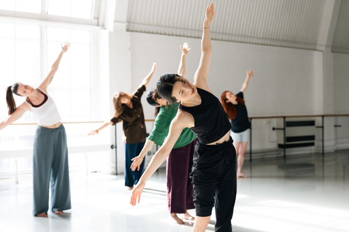 People performing a dance or exercise routine in a sunlit studio, with arms extended and leaning to one side.