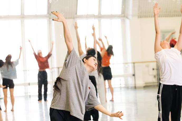 A group of people in a dance studio stretching with arms raised, with sunlight streaming through large windows in the background.