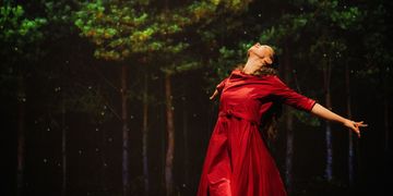 A dancer in a flowing red dress dances gracefully in a forest at night, with trees and small lights in the background.