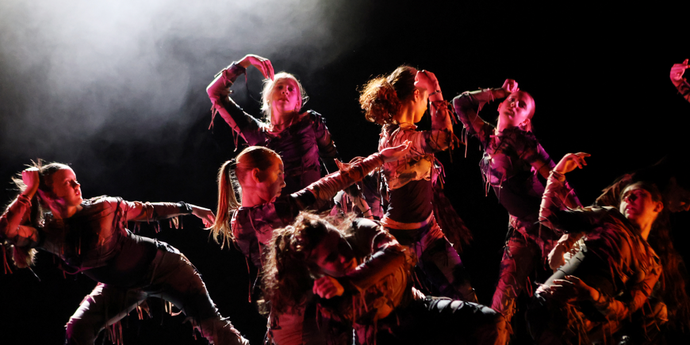 Dancers in tattered costumes perform expressively on stage, illuminated by dramatic lighting and surrounded by mist.