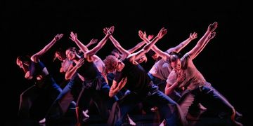 A group of dancers perform on stage, striking expressive poses with outstretched arms in a dynamic, dimly lit setting.