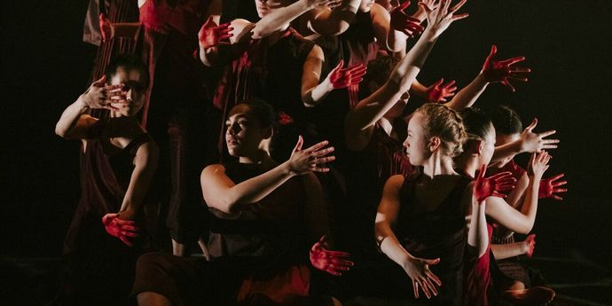 A group of dancers in dark outfits perform under dramatic lighting, with red-painted hands creating intricate shapes and movements.