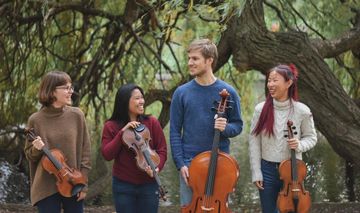 Four people outdoors, smiling while holding string instruments: two violins, a viola, and a cello, with trees in the background.