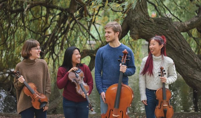 Four people outdoors, smiling while holding string instruments: two violins, a viola, and a cello, with trees in the background.