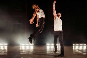 Two dancers in white tank tops and black pants perform energetically on stage, one leaping high while the other extends an arm upward.