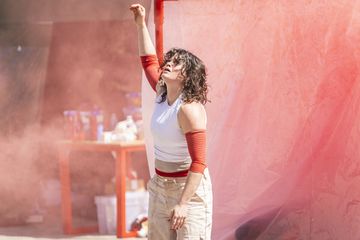 woman with hand in air wearing white top and beige trousers. dust in the air against a red sheet background