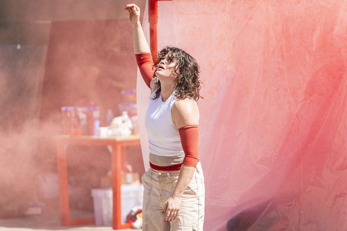 woman with hand in air wearing white top and beige trousers. dust in the air against a red sheet background