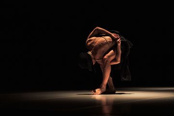 A dancer in a dimly lit space performs a contorted pose, showcasing muscular back and arms, with dramatic shadows on the floor.