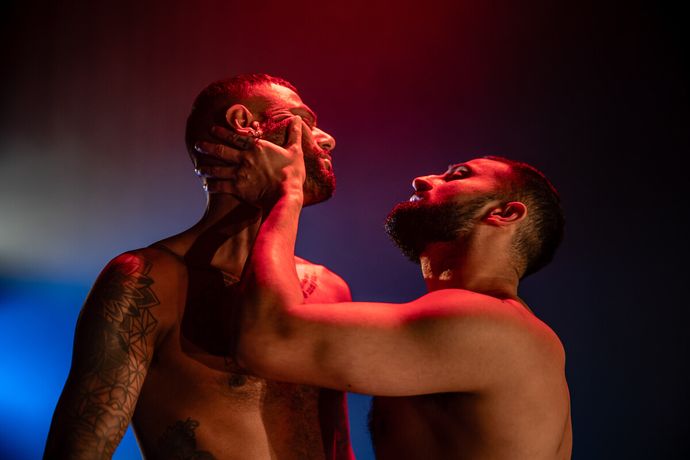Two shirtless individuals under dramatic lighting; one gently holds the other's face, creating an intense, expressive scene.