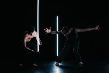 Two dancers perform in dim light, each interacting with vertical light beams, creating a dramatic and dynamic scene on stage.