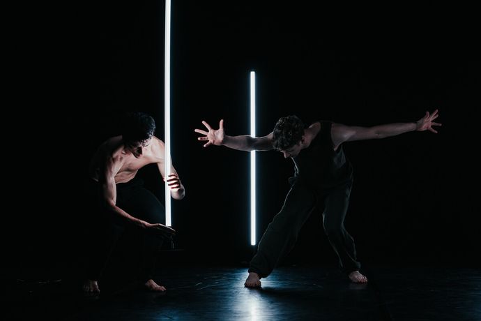 Two dancers perform in dim light, each interacting with vertical light beams, creating a dramatic and dynamic scene on stage.
