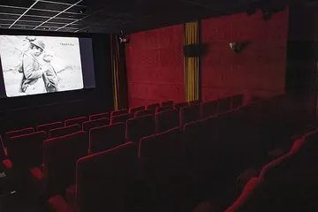 Empty movie theater with red seats, showing a black-and-white film on screen. The room is dimly lit.