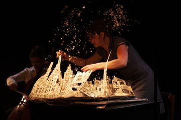 A woman performs with a paper model of a city amid flying debris on stage, while a cellist plays in the background in dim lighting.