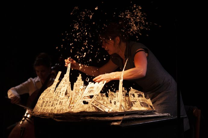 A woman performs with a paper model of a city amid flying debris on stage, while a cellist plays in the background in dim lighting.