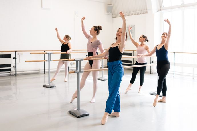 Ballet dancers in a studio practicing at the barre, each performing a pose with one arm raised. Bright, airy space with large windows.
