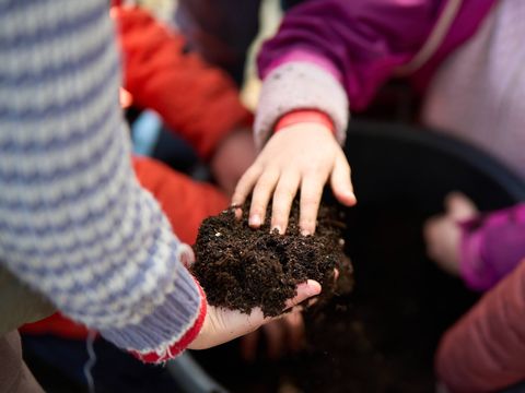 Children in patterned jumpers and jackets using their hands to handle soil above a large container.
