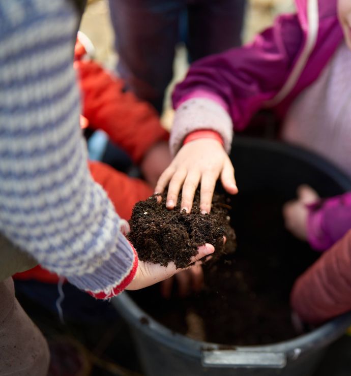 Children in patterned jumpers and jackets using their hands to handle soil above a large container.