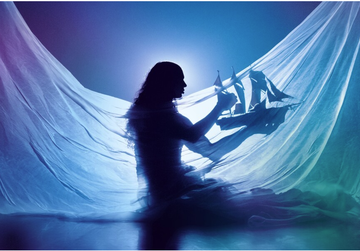 Silhouette of a person holding a model ship, surrounded by flowing fabric, with a blue and purple backlight creating a dreamy atmosphere.