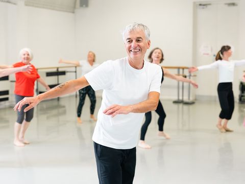 A group of older adults, led by a smiling man in a white shirt, participates in a dance class, each with arms extended, in a bright studio.
