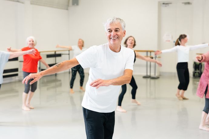 A group of older adults, led by a smiling man in a white shirt, participates in a dance class, each with arms extended, in a bright studio.