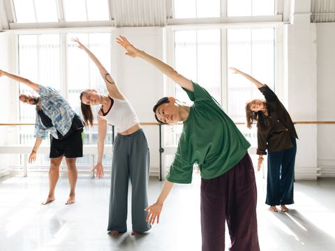 A group of four people stretches with arms overhead in a brightly lit dance studio, bending sideways in unison.