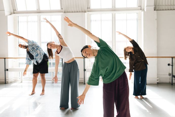 A group of four people stretches with arms overhead in a brightly lit dance studio, bending sideways in unison.