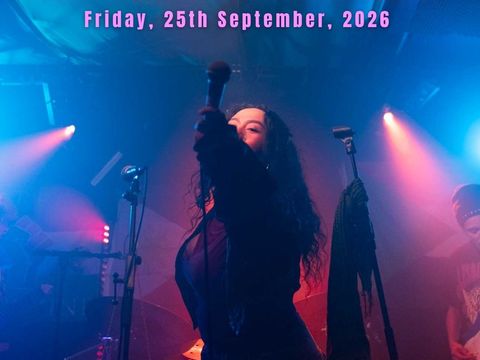 Person with long curly hair holding a microphone on a stage with colourful lights, standing in front of musicians and musical equipment.