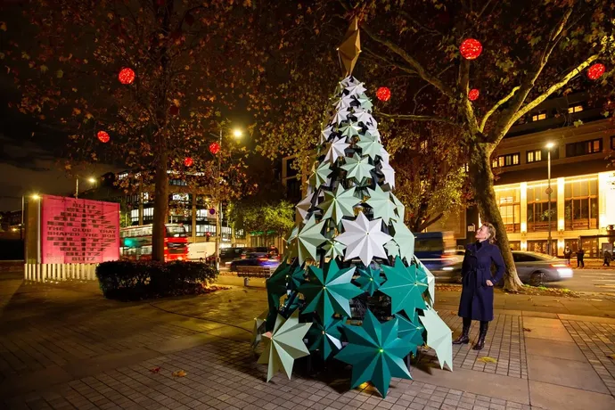 A large outdoor Christmas tree sculpture made of geometric star shapes in green and white, illuminated on a city street at night.