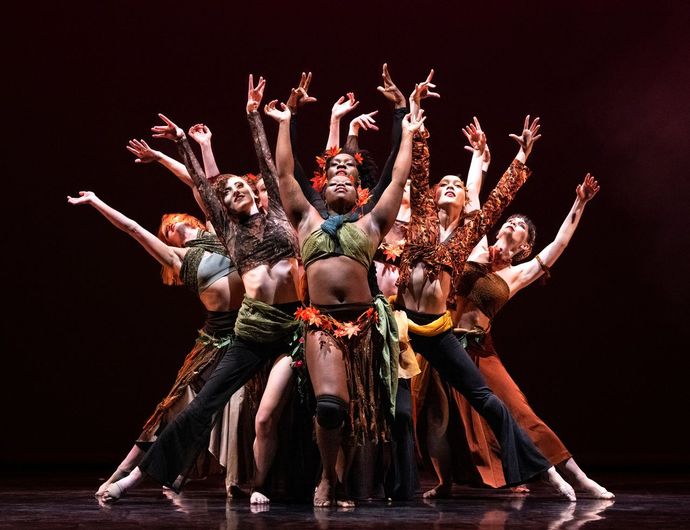 A group of dancers in colorful costumes perform a dynamic routine on stage, with arms raised expressively against a dark background.