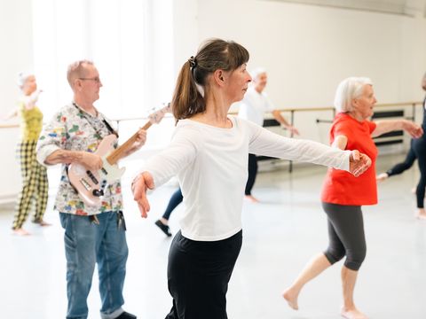 A group of mature adults participates in a dance class, while a man in the background plays an electric guitar.