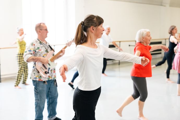 A group of mature adults participates in a dance class, while a man in the background plays an electric guitar.