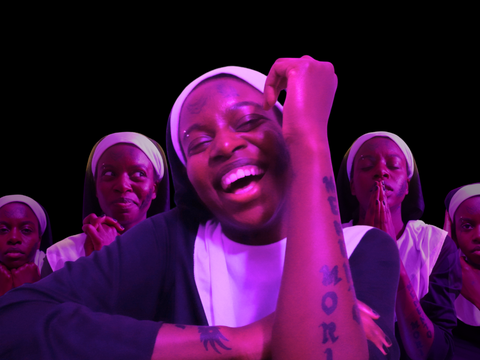 Person dressed in a nun's habit with visible tattoos on their face and arm, smiling with other similarly dressed figures in varied poses against a black background.